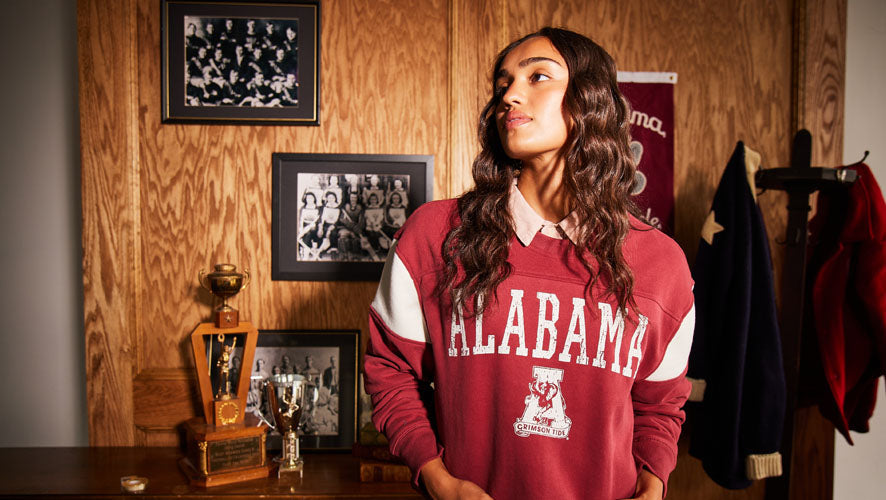 Female model in a crimson Alabama sweatshirt with bold varsity lettering from ‘47 Brand, standing next to trophies and retro photos in a wood-paneled room.