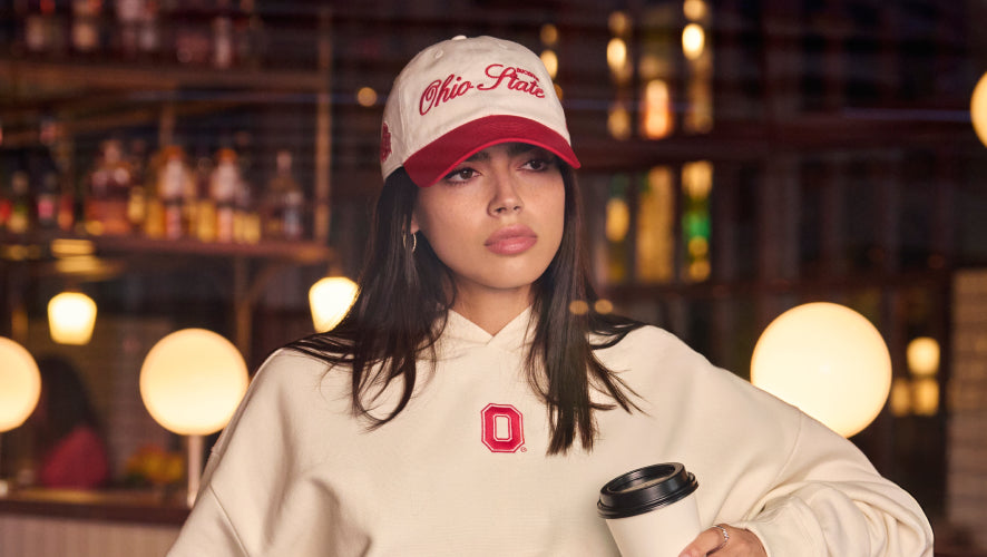 Person wearing Ohio State-themed hoodie and cap holding a coffee cup in a bar setting