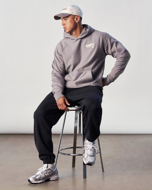 Person wearing a gray hoodie with a logo, black pants, and white sneakers, sitting on a stool against a plain background.