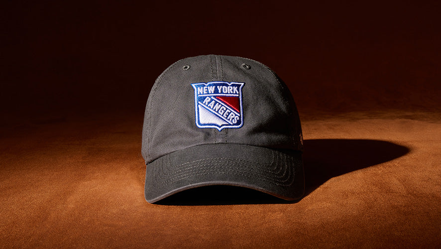 Black New York Rangers hat with embroidered shield logo, placed on a warm brown surface with dramatic lighting.
