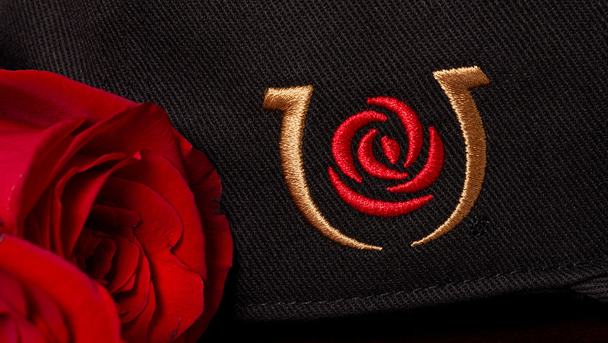 A detail image of a horseshoe logo with a red rose on the inside embroidered on a black hat. There is a red rose next to the hat.