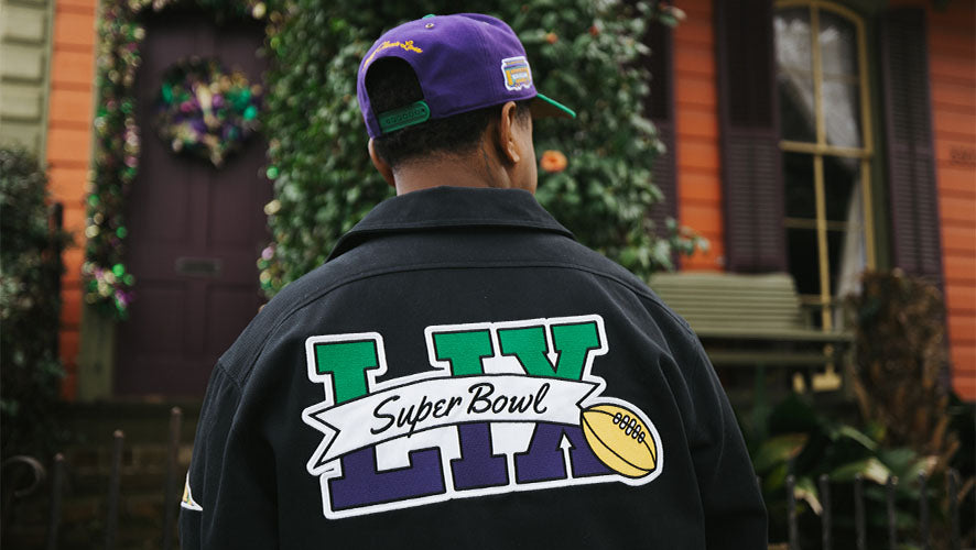 Man wearing a black Super Bowl LIX jacket and purple Mardi Gras-themed cap, standing outdoors in front of a decorated New Orleans-style house.
