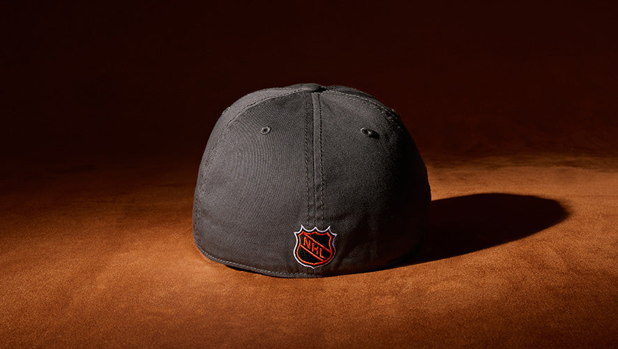 Rear view of a dark grey cap featuring a small red and black NHL shield logo, set against a dramatic brown background.