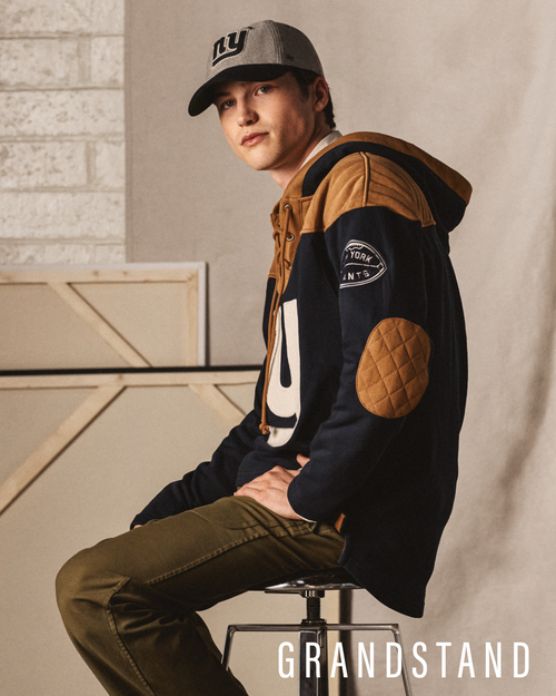 Person wearing a sweatshirt with patches and a cap, sitting on a stool against a neutral background. GRANDSTAND