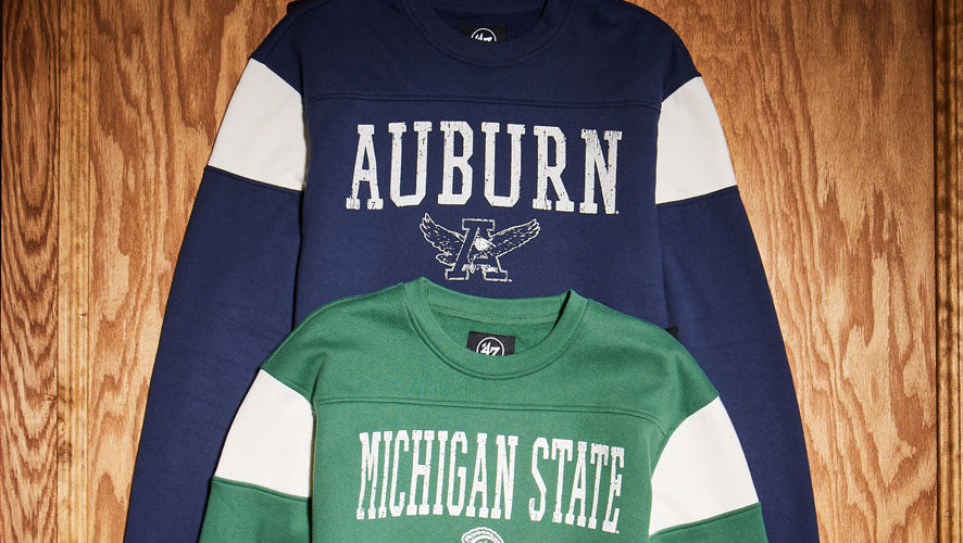 Flat lay of two retro collegiate crewnecks from ‘47 Brand featuring Auburn and Michigan State designs, with bold vintage lettering on striped sleeves.