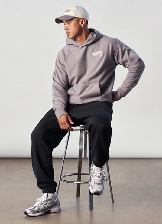 Person wearing a gray hoodie and black pants sitting on a stool against a white wall.