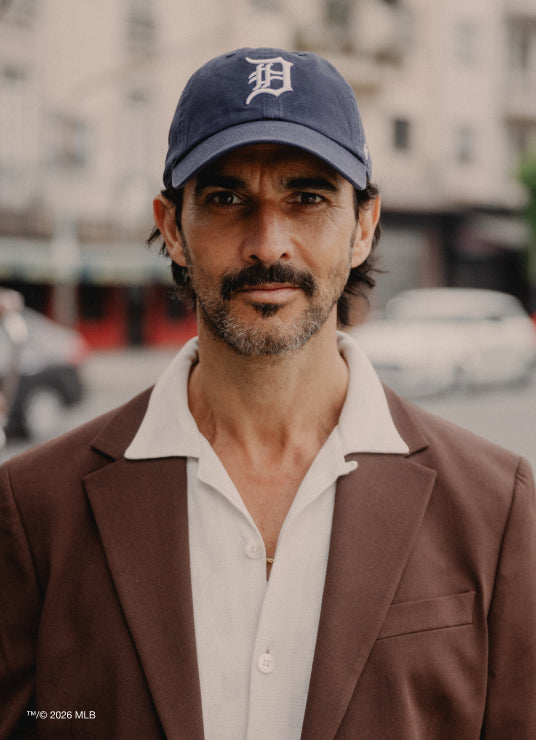 Man wearing a blue cap with a logo and brown jacket in an urban setting