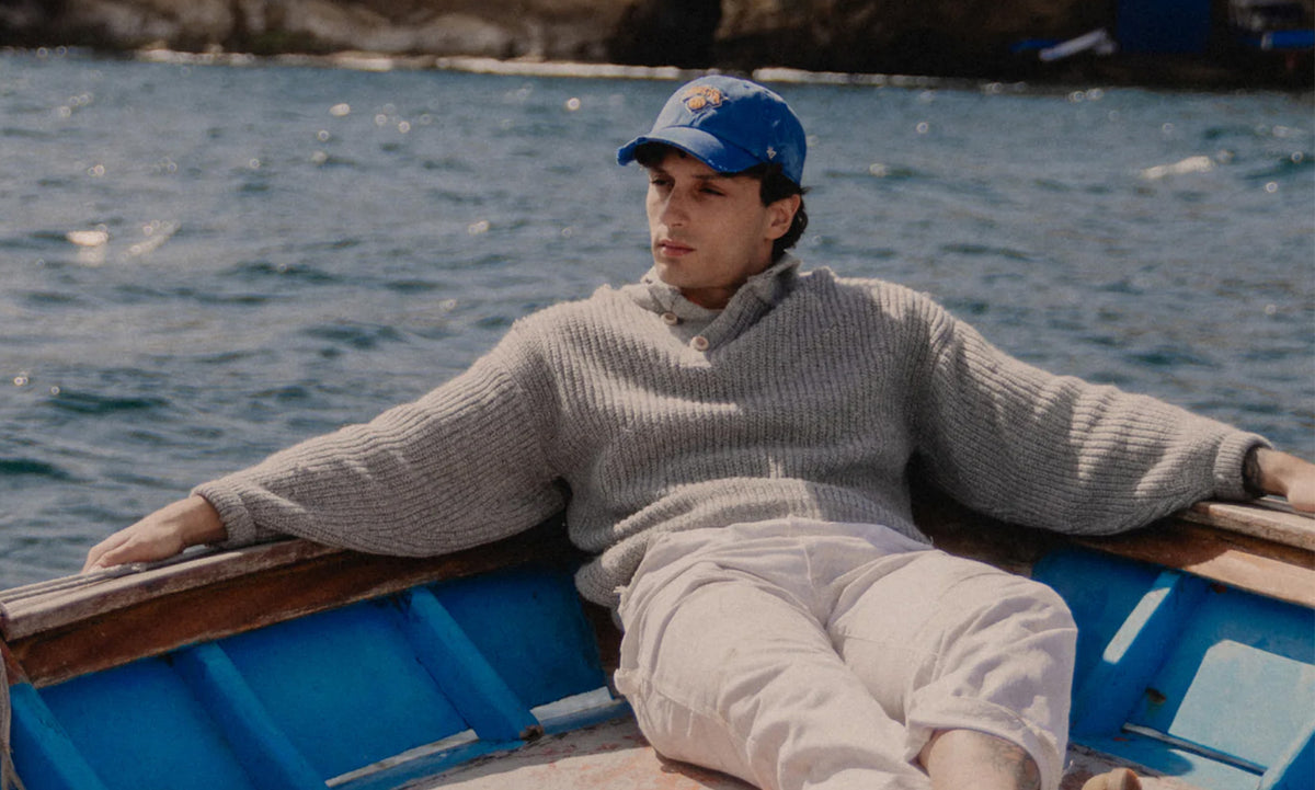 Person sitting in a small boat on a body of water wearing a cap and sweater.