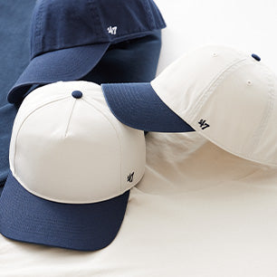 Minimalist ‘47 Brand hats in white and navy, including baseball caps and dad hats, displayed on a soft cream backdrop.
