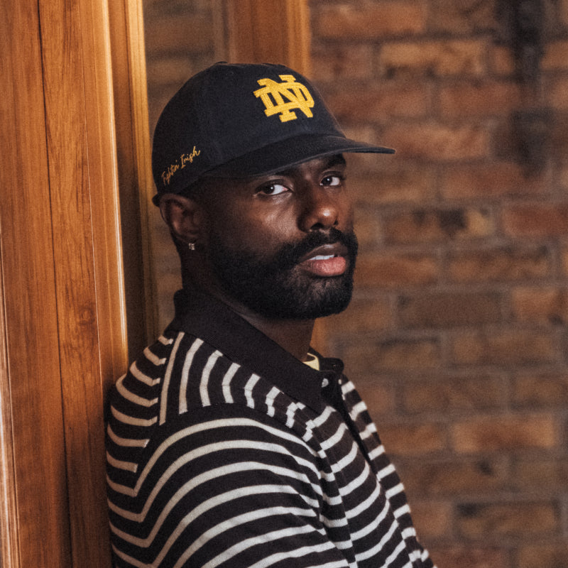 Man wearing a black cap with a logo and a striped shirt against a brick wall.