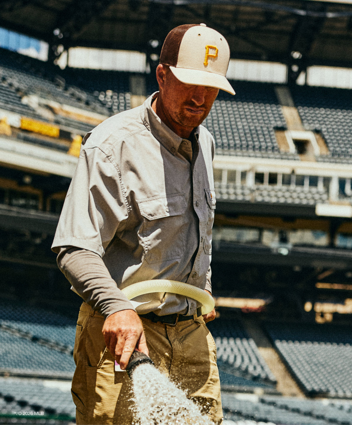 Man in a baseball cap and work clothes at a stadium, holding a hose.