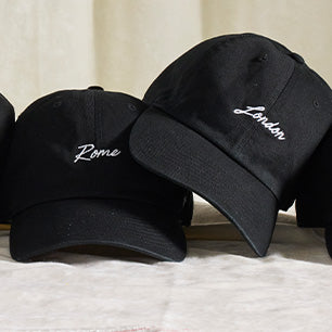 Black embroidered baseball caps from ‘47 Brand featuring sleek white script city names like Rome and London, displayed on a soft surface.