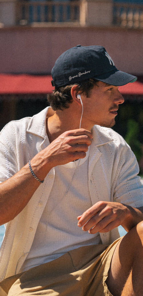 Man wearing a navy cap and white shirt, sitting outdoors with earphones.