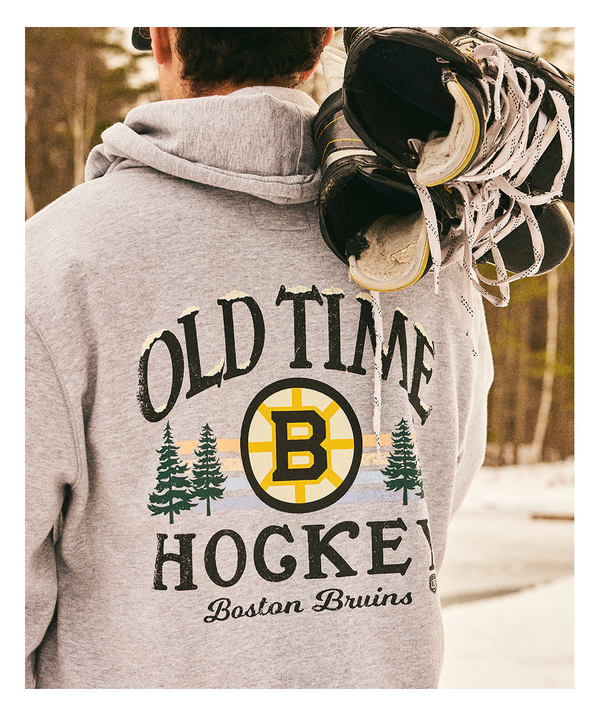 Person wearing a gray 'Old Time Hockey' Boston Bruins hoodie with hockey skates on their shoulders, set against a snowy background.
