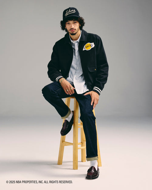 Person wearing a black jacket with a logo, sitting on a stool against a gray background.