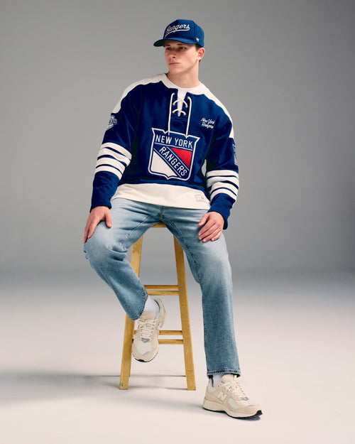 Person wearing a New York Rangers sweater and cap sitting on a stool against a gray background