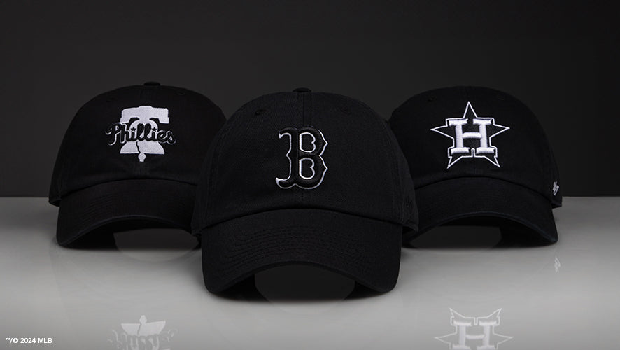 Three black caps with white MLB team logos (Phillies, Red Sox, Astros) arranged in a row on a reflective surface.