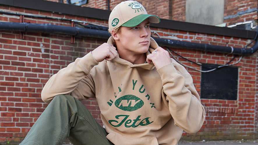 Male model wearing a tan hoodie with green “Jets” text and a matching beige and green cap, sitting outdoors against a brick wall.