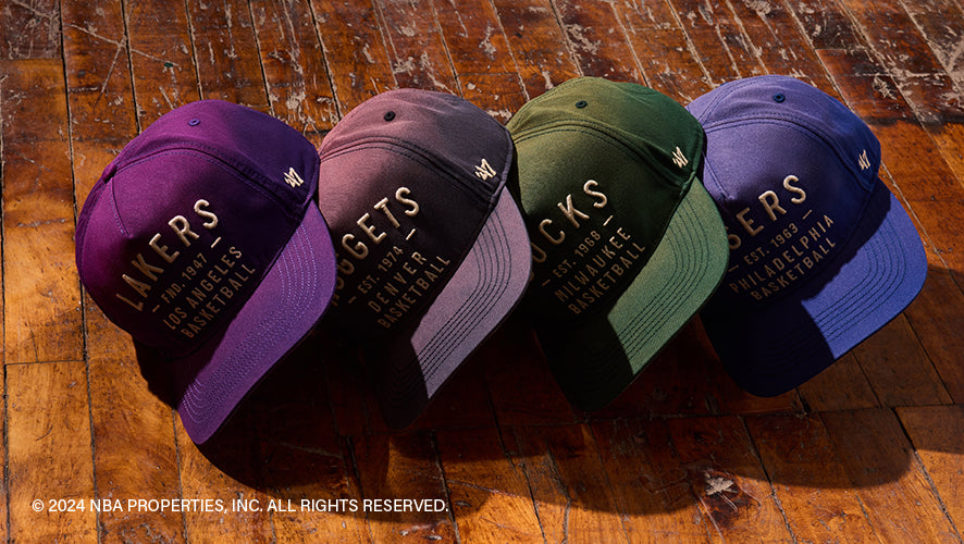 Four two-tone NBA hats laid on wooden floor, each with bold script logos for Lakers, Nuggets, Bucks, and Sixers in matching tonal colors.