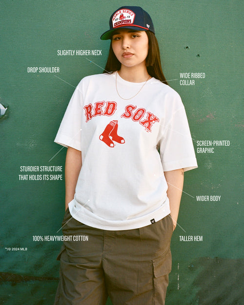 Woman wearing a Red Sox tee and World Series cap with labeled features including drop shoulder, screen-printed graphic, slightly higher neck, sturdier structure that holds its shape, heavyweight cotton, wider body, taller hem, and wide ribbed collar.