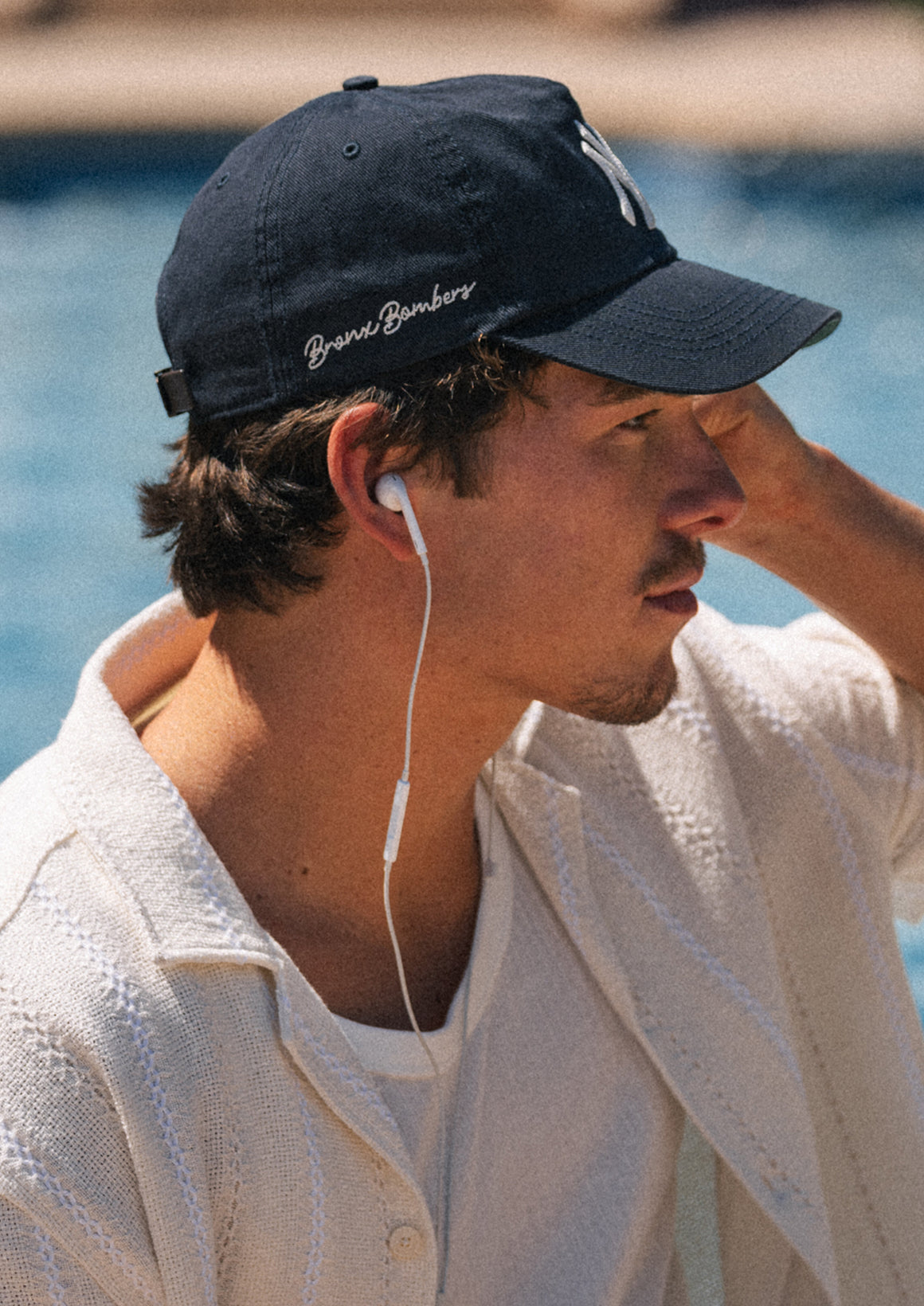 Man wearing a navy cap with 'Brixx Bomber' logo, listening to music with earphones, by a body of water.