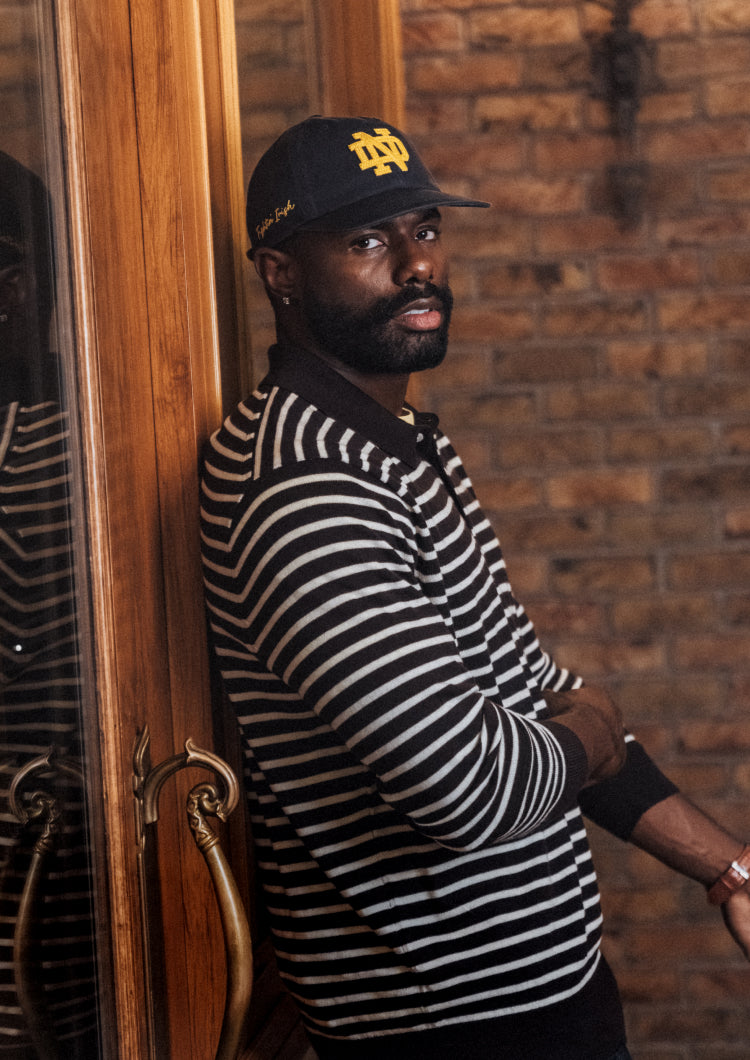 Man wearing a striped shirt and cap leaning against a wooden door with a brick wall background