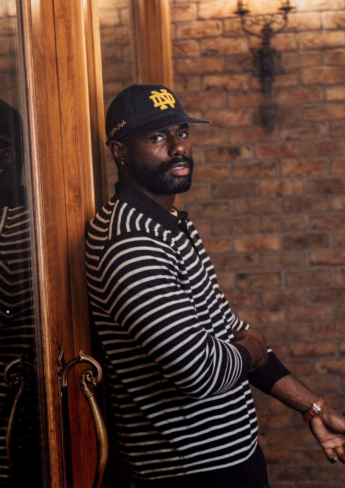 Man wearing a striped shirt and cap leaning against a wooden door with a brick wall background