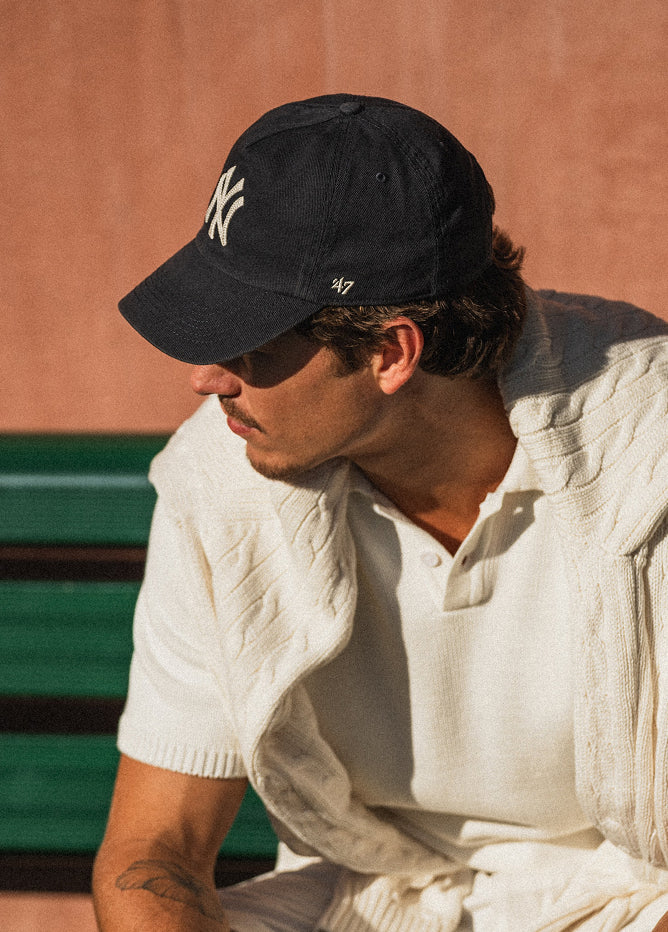 Man wearing a black cap with a logo, sitting on a green bench against a brown wall.