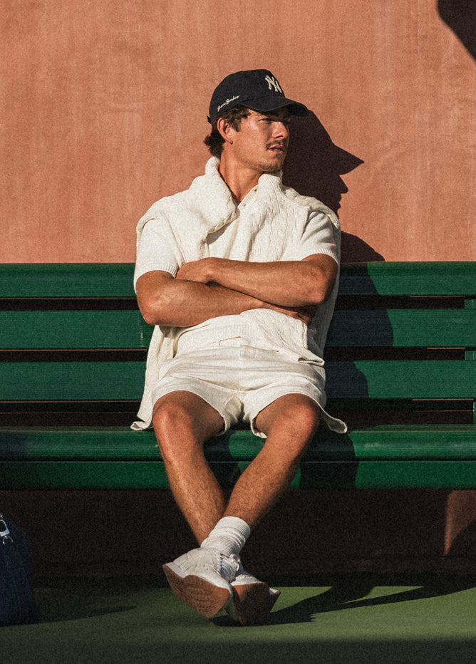 Person sitting on a green bench with a sweater draped over their shoulders, wearing a cap and white outfit.