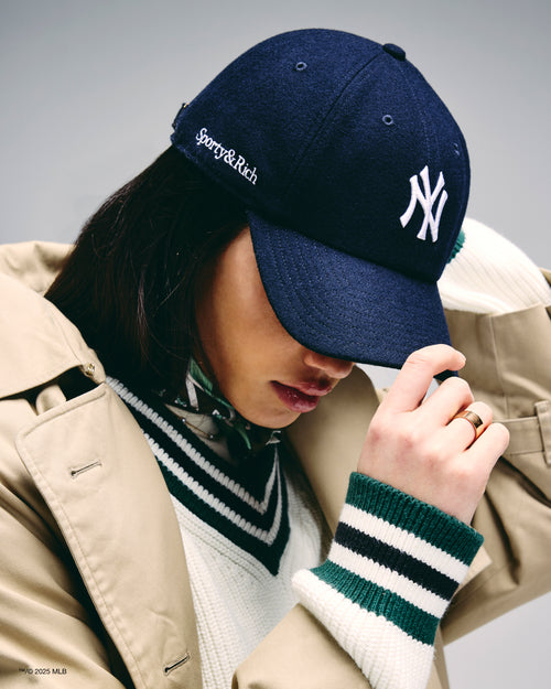 Person wearing a navy blue cap with a logo, beige coat, and striped wristband on a gray background