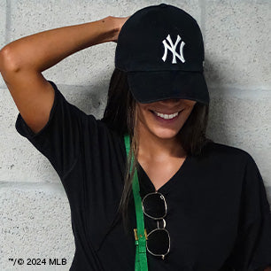 Woman in a black Yankees cap and shirt smiles with one arm raised, posing against a concrete wall.