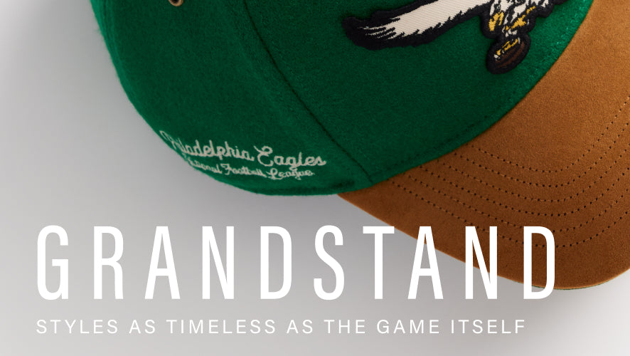 Two baseball caps, one green and one brown, on a light gray background with 'Grandstand' branding. GRANDSTAND- styles as timeless as the game itself