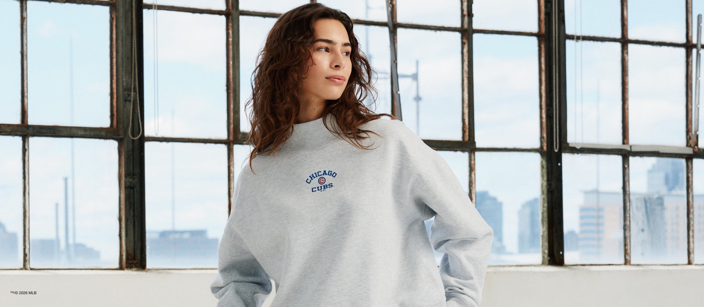 Woman wearing a light gray sweatshirt with a logo in front of large windows with a cityscape view.
